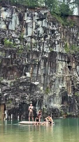 A video shows four people on a raft in a quarry lake. A steep, rocky cliff face dominates the background. The people are positioned around the raft, with two standing and two sitting. The water is calm and reflects the cliff.