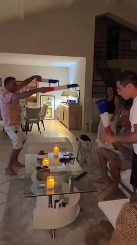 In a dimly lit living room, a group of people are gathered. A man in a patterned shirt and white shorts stands in the center, enthusiastically waving two French flags. He is facing a man sitting on a couch who is also holding a French flag. To the right, another man in a white t-shirt is observing the scene, and behind him, a woman is watching. In the foreground, a glass-topped coffee table holds lit candles and various items. The room features a fireplace, wooden cabinets, and artwork on the walls, suggesting a home environment. A dog is visible near the couch on the left. The atmosphere appears festive and celebratory.