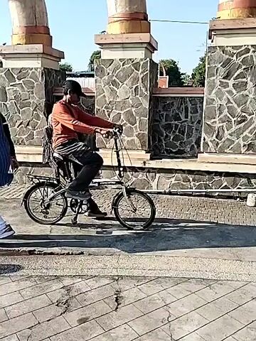 A medium shot from ground level shows a man in a red long-sleeved shirt and a black baseball cap pushing a white folding bicycle past a stone and concrete wall. He stops next to a yellow trash can and a white pole. In the background, there are people standing and walking under a blue canopy and trees. The ground is paved with square tiles. The weather appears sunny.