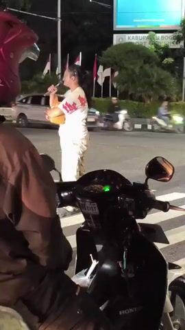 The image captures a nighttime street scene in Cibinong, Indonesia. In the center, a woman stands on what appears to be a zebra crossing or pedestrian area, holding a microphone and a brown folder or book. She is wearing a white t-shirt with a design and patterned pants, and her hair is pulled back. She appears to be singing or speaking into the microphone, possibly street performing.

In the immediate foreground, the handlebars and dashboard of a black Honda motorcycle are visible, with a person partially obscured behind them, suggesting they are stopped. A sticker on the motorcycle's dashboard reads "RACIST," and "HONDA" is visible on the motorcycle's body.

The background features a busy road with blurry car lights, indicating moving traffic. Streetlights illuminate the scene. To the right, a large billboard displays a blue advertisement with the text "www.ntmmedia.co.id." Below the billboard, a sign is visible with text "KABUPATEN BOGOR SPORT & TOURISM." Several flags, possibly Indonesian flags, are lined up behind the sign. The overall ambiance is that of an urban area at night.
