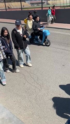 The video shows a street scene in China on a sunny day. Three young women walk across the street in the foreground. They are dressed casually in jeans and jackets, and two of them wear baseball caps. One carries a water bottle. Behind them, people are walking on the sidewalk and riding electric scooters in the street. In the background, there is a large white building, likely a school or hospital, behind a fence. The sky is blue with some clouds. The camera moves slightly during the recording.