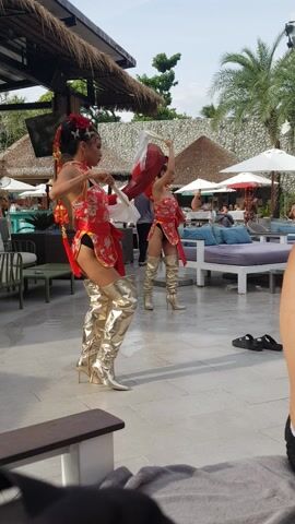 Two performers, possibly drag queens, are engaged in a dance routine on a patio. They both wear elaborately styled hair with floral decorations, red and white cheongsam-style dresses, and high-heeled gold boots. The performer in the foreground holds a white fan in one hand, while the performer behind her holds a red scarf. The dance is taking place in an outdoor setting, possibly a resort, with thatched-roof structures, palm trees, white umbrellas, and lounge seating in the background. Several people are sitting on the lounge furniture, possibly watching the performance. The flooring is tiled. The video is vertically oriented and includes a partial view of a person's leg and shoe in the upper right corner.
