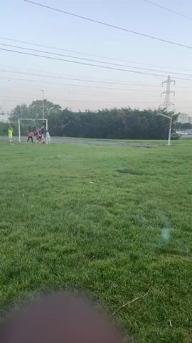 The video shows a grassy field with a soccer goal at one end. Several people are gathered near the goal. There is a line of trees and bushes behind the goal. In the background, there are power lines and a tower. The sky is clear and the light suggests it is either early morning or late afternoon. There is some blur or obstruction in the lower foreground of the video, possibly from a finger or object near the camera lens.