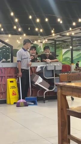 The video shows a cleaning crew with a broom, dustpan, and wet floor sign in what appears to be an indoor dining area with tables. Two young men, likely workers, are leaning over a counter, one holding a broom and the other a dustpan, as a third person in a uniform, also wearing a face mask, stands and talks to them. String lights are visible overhead.