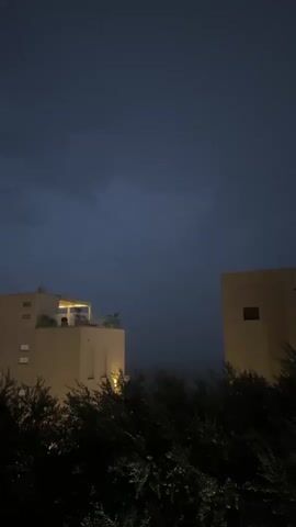 This media file captures an outdoor scene at dusk or night in Trap, Montenegro. The primary subjects are two modern, flat-roofed buildings with light-colored facades, one on the left and one on the right of the frame, separated by a dense area of dark green foliage. The building on the left has multiple windows and a lit rooftop terrace. The building on the right has a single visible window. The foliage in the foreground obscures a clear view of the ground level, but suggests a residential or resort-like setting, with two parked cars visible towards the bottom right.

The sky is a deep, dark blue, indicative of evening or stormy weather. There are subtle hints of lightning in the distant sky above the buildings, suggesting a thunderstorm is occurring or approaching. The lighting is dim, with some artificial lights illuminating parts of the buildings and the foliage, and a streetlamp is visible casting light on the parked cars.

The overall atmosphere is moody and slightly dramatic, due to the dark sky and implied storm. The absence of people suggests a quiet moment, perhaps people are indoors seeking shelter from the weather. No visible text is present in the media file.