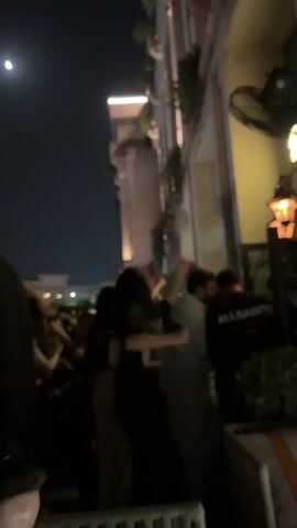 A medium shot of an outdoor night event shows several people dancing and mingling in front of a building with plants and lights. On the left side of the frame, a bright full moon is visible in the dark sky. The building has a cream-colored facade with several columns and balconies adorned with hanging green plants and small pink flowers. Bright lights illuminate the top floor of the building. On the right side of the frame, an outdoor wall-mounted fan is visible in the upper right corner, and a decorative gas lantern in the lower right corner, casting a warm glow on the wall. The people in the video are mostly young adults. In the foreground, two women are dancing facing away from the camera, one wearing a black top and light-colored pants, and the other wearing a black dress and carrying a white handbag. Behind them, a man in a light green shirt and a man in a black long-sleeved shirt with "ALLSAINTS" written on the back are seen talking and laughing. The atmosphere appears lively and festive, with music playing in the background.