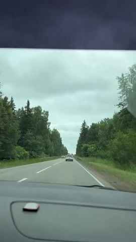 The video shows a view from inside a car driving on a paved road surrounded by trees. Several cars are visible on the road ahead. The sky is cloudy. The dashboard of the car is visible in the foreground.