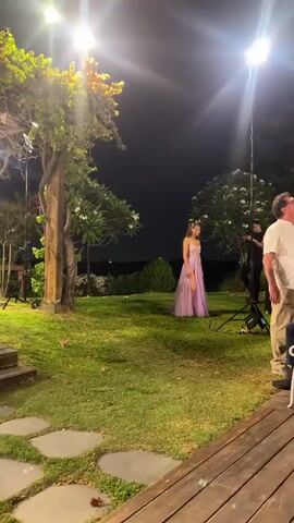 A young woman in a long, light purple dress with a high slit and a plunging neckline poses for a photograph outdoors at night. She is standing on a grassy lawn, with trees and foliage in the background. A man is standing behind her to the right, holding a camera and seemingly taking her picture. A tall light stand is visible to the right of the man. There is a wooden post structure to the left of the frame, partially obscured by a tree with illuminated foliage. The scene is lit by artificial lights, creating a dramatic effect.
