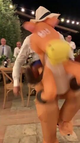 A person in an inflatable horse costume is walking past a seated audience at an outdoor event at night. The audience members are seated at tables and appear to be clapping and smiling. String lights are visible overhead. The person in the costume is wearing a white shirt and a cowboy hat. The costume is orange and brown and has a yellow bell on the horse's neck.