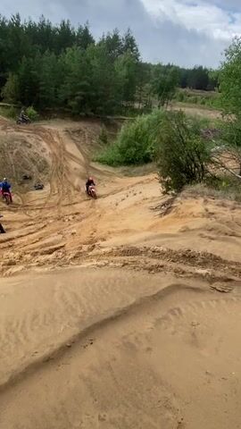 A group of people are dirt biking on a sandy track. One person on a red dirt bike is seen going down a sandy hill. As they reach the bottom, the dirt bike slides and the rider falls off. The rider is wearing a red helmet and black and red riding gear. There are other dirt bikes and riders visible in the background. The environment is a sandy area with trees and green foliage.