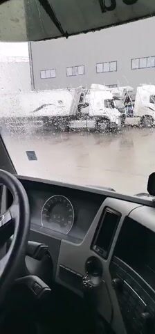 The video is shot from inside the cabin of a vehicle, likely a truck, as evidenced by the dashboard, steering wheel on the left, and visible windshield wipers. Heavy rain is pouring down, visible through the water droplets on the windshield and the wet ground outside.

The vehicle appears to be moving slowly or turning within an industrial area. Outside, several white semi-trucks are parked or waiting. One truck in the foreground, seen through the rain-streaked windshield, has a large white bulk trailer with "WOLTON" clearly visible on its side. In the background, there are gray industrial buildings with rectangular windows. As the vehicle pans, more industrial structures become visible, including scaffolding, large pipes, and what appears to be part of a processing plant. The ground is uniformly wet, reflecting the gray, overcast sky.

The scene depicts an industrial yard during a rainy day in Karapelit, Bulgaria. No people are directly visible, and the focus is on the industrial environment and the challenging weather conditions.