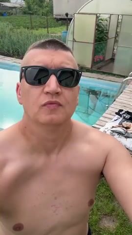 A shirtless man with short hair and sunglasses stands in front of a swimming pool and a greenhouse. He looks at the camera, then smiles and waves.
