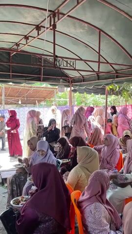 The video shows an outdoor gathering of people, primarily women, seated under a green canopy supported by a metal frame. The individuals are mostly wearing headscarves and traditional attire in various colors, predominantly shades of pink, purple, and beige. Many are seated on orange plastic chairs, while others are seated on the floor on patterned fabric. Some people are holding plates and appear to be eating, with food visible on the plates. There are also some children present, and a few men are visible in the background. The environment appears to be a casual event, possibly a celebration or a communal meal, with foliage and trees visible in the background. The lighting suggests it is daytime.