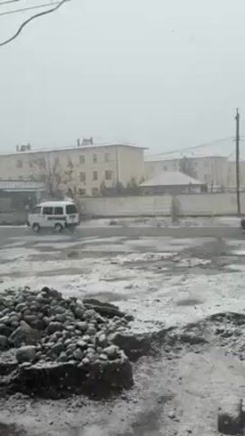 The video shows a view of a snowy, urban area. The ground is partially covered in snow with patches of visible asphalt or dirt, and a pile of dark rocks sits in the foreground. Across the road, there are a few light-colored buildings with flat roofs, some with a reddish tint from snow accumulation. A light gray wall runs in front of the buildings. Power lines are visible in the sky. Two vehicles drive by: a dark-colored SUV and a white van with red stripes. The sky is overcast and snow is falling.
