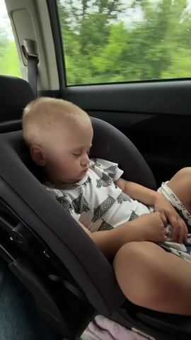 A blond-haired toddler is asleep in a car seat in the backseat of a car. The toddler is wearing a white t-shirt with a black and white pattern of what appears to be cats or other animals and has light-colored shorts. The car seat is dark gray. The background shows a blurred view of green trees through the car window, indicating the car is in motion. The lighting suggests it is daytime.