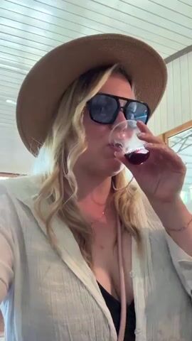 A close-up video shows a woman, likely at a wine tasting or similar event, drinking from a small cup and listening intently. She has long, wavy blonde hair and wears a wide-brimmed straw hat. Dark sunglasses with a pink beaded chain rest on her nose. She is dressed in a loose-fitting white textured linen shirt worn open over a dark top, revealing a delicate gold-toned necklace. A subtle bracelet is visible on her right wrist.

In the initial frames, she is seen taking a sip of a dark red beverage from a small, clear plastic tasting cup, her eyes seemingly closed in enjoyment or contemplation. She then lowers the cup and looks to her right, as if listening intently to someone speaking off-camera.

The setting appears to be indoors, possibly a rustic yet bright space with a white-slatted wooden ceiling. In the background, what looks like a large window or doorway allows natural light to enter, reflecting off surfaces and revealing hints of an exterior structure. The atmosphere seems relaxed and informal, typical of a casual daytime gathering.

The accompanying audio features a woman speaking in Russian, discussing the positive effects of something on the skin and its rejuvenating properties, suggesting she might be listening to a presentation or commentary during the event. The location context provided is Rassvet, Russia.