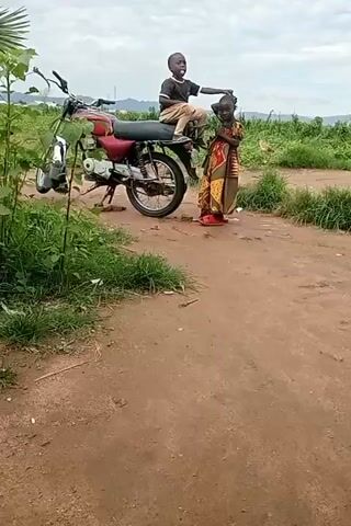 A young boy is sitting on the back of a red motorcycle. A young girl is standing next to the motorcycle. The girl is wearing a colorful dress and red shoes. She is holding a piece of fabric. The boy is wearing a black shirt and light-colored pants. He is looking at the camera and smiling. The girl is looking at the camera and smiling. The motorcycle is parked on a dirt road. There is green vegetation on the left side of the road and in the background. The sky is cloudy.