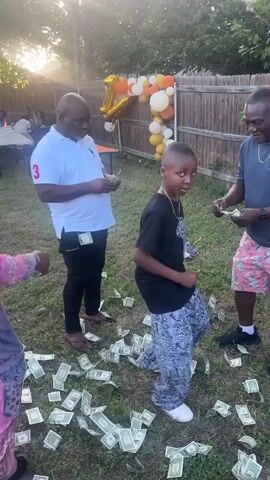 A sunny backyard birthday party scene with a young Black boy standing amidst a scattered pile of dollar bills. He’s dressed in a black T-shirt, patterned pants, and white shoes.  Three adults surround him, two handing him money and one embracing him. To the boy’s left, another child is seen picking up money. A balloon arrangement with a large number “1” is visible in the background. A wooden fence surrounds the yard, and more guests are visible at a table on the left.