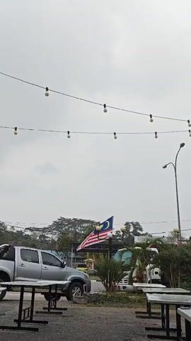 This video shows an outdoor scene with a cloudy sky. There are two strings of lights hanging across the frame. In the foreground, there are several empty tables and benches. To the left, a silver pickup truck is parked. In the middle ground, a Malaysian flag is flying on a pole. Behind the flag, there is a white truck and various greenery. A lamppost is visible on the right side of the frame. A sign with a logo and text is mounted on a pole on the right.
