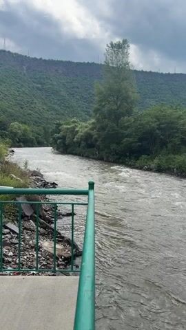 A video shows a river flowing through a green, mountainous landscape under a cloudy sky. A green railing is visible in the foreground, and on the left side of the river, there appears to be some debris and a small building. The river is wide and appears to be flowing with some force, creating ripples on its surface. The surrounding hills are covered in lush green trees and vegetation. The sky is overcast with dramatic clouds, suggesting the possibility of rain.