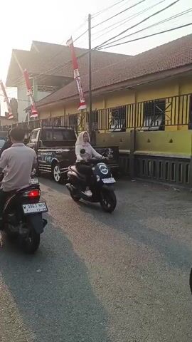 The video shows a street scene with multiple motorcycles and a few cars. The sun is setting, casting a warm light on the scene. There are people riding on the motorcycles, most of them wearing helmets. Some of the motorcycles are carrying more than one person. Red and white flags are visible on poles along the street, suggesting a festive or celebratory occasion. The buildings on the side of the street appear to be commercial or residential. The road is paved and there are power lines overhead. The overall impression is of a busy street with traffic moving in the same direction.