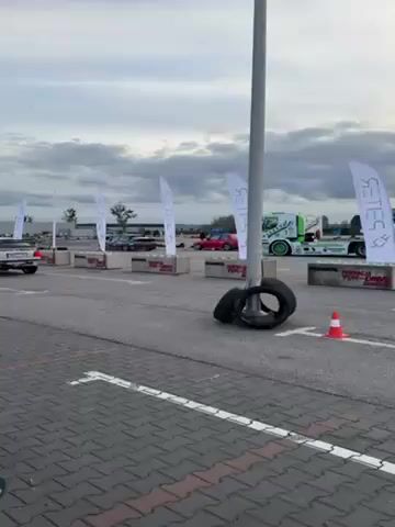 The video shows a parking lot that has been set up for a racing event. White flags with a logo on them line the side of the track, which is marked with white lines. There are concrete barriers on the sides of the track, and two cars can be seen in the distance, one red and one dark colored. A green tow truck is parked on the side of the track, and a stack of tires and a traffic cone are visible in the foreground. The sky is overcast, and the ground is paved with bricks. A person is recording the video on a phone. A car passes by at the start of the video.