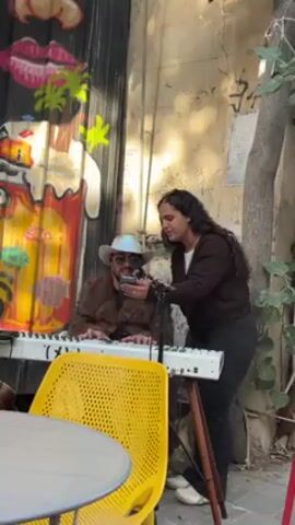 A woman sings into a microphone as a man plays a keyboard in front of a wall covered with graffiti. She stands, facing forward and slightly to the left, dressed in a dark brown long-sleeved shirt and black pants. She has long, dark, curly hair. The man sits on a chair behind a white keyboard, wearing a brown furry jacket, sunglasses, and a cowboy hat. He's looking down at a smartphone he holds near the microphone. A yellow plastic chair sits next to a round white table in the foreground. The wall behind them has an illustrated, colorful graffiti mural. Next to the wall is a tree with green leaves and a weathered bark. The scene is set outdoors in daylight.