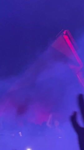 The video shows a dimly lit environment with a fog machine, creating a hazy atmosphere. A large, geometric structure, possibly part of a stage setup, dominates the view. Laser lights, shifting between red and green, cut through the fog, highlighting the structure. People are visible in the lower part of the frame, seemingly enjoying a performance or event.