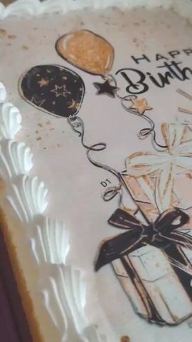 The video shows a close-up, overhead view of a cake. The cake is decorated with an edible image featuring balloons, gift boxes, and the words "Happy Birthday." The cake has white frosting around the edges. The lighting is bright, and the focus shifts slightly throughout the clip, providing different perspectives on the cake's design.