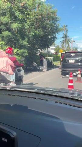 The video shows a road scene on a sunny day. On the left side of the road, two people are visible on a scooter, one driving and one passenger wearing a pink hoodie. The scooter appears to be parked or stopped. In the middle of the road, a black SUV is parked, with its rear facing the camera. Two orange traffic cones are placed in front of the SUV, suggesting some sort of obstruction or hazard. Further down the road, another scooter passes by, ridden by a person wearing a blue shirt. The environment appears to be somewhat rural or suburban, with trees and power lines visible. The perspective is from inside a vehicle, looking out through the windshield.
