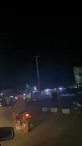 The video shows a street scene at night. The location appears to be a commercial area with small shops and stalls illuminated by streetlights and the lights of the shops.  People are walking around and cars and motorcycles are driving on the road. In the beginning of the video, a person is riding a motorcycle in front of the camera. A decorative, lit up object, possibly a small Ferris wheel, is visible in the distance.