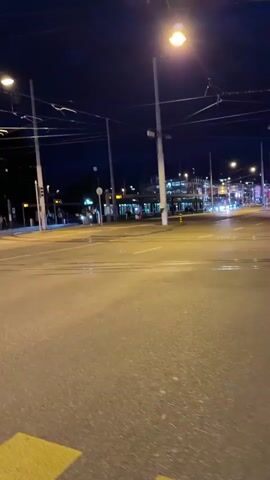 The video shows a dark street at night with several streetlights illuminating the area. A two-story building with a yellow facade is visible on the left side of the street. In the foreground, there is a yellow pedestrian crossing. A green and gray bus drives from left to right across the screen, passing the building and the pedestrian crossing.