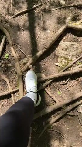 The video begins with a close-up shot of the ground, covered in dirt and exposed tree roots. A person's foot in a white sneaker appears in the frame. The camera then pans up to reveal a clear, shallow stream flowing over rocks covered in moss. The surrounding environment consists of a dense forest with trees lining the stream. Sunlight filters through the canopy, creating a dappled light effect on the water and surrounding vegetation. The stream appears to be relatively undisturbed, flowing peacefully through the woodland.