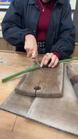 A woman stands at a wooden table and cuts a long green plant stalk in half using a knife and wooden cutting board. The stalk and cutting board are on top of a light brown or beige cloth. To the right of the cutting board is a wooden rolling pin. The woman is wearing a black coat over a red turtleneck sweater, black belt, gold rings and a necklace with an A pendant. After cutting the stalk, she picks up the two pieces and talks to someone off screen. She then turns away from the table toward a wall covered in framed art depicting Ancient Egyptian artwork. The table in front of the wall contains a yellow mallet and blue trays. She points to the wall and appears to be explaining the art.