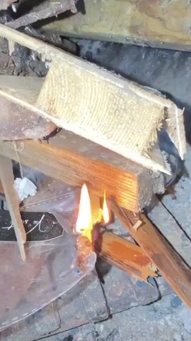A small fire is burning among pieces of wood and what appears to be a clear plastic object. Flames are visible consuming the plastic and igniting the wood. The environment seems to be outdoors or in a structure with exposed wood and rough surfaces. The lighting is dim, and the focus is on the burning materials.