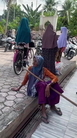 The video captures various moments of people gathering outdoors in Meuke Beurabo, Indonesia, likely at a community or religious site.

The first shot focuses on an elderly woman sitting on the edge of a raised concrete platform, resting on a paved area with hexagonal tiles. She wears a blue hijab, an orange long-sleeved top, and a purple sarong, and holds a wooden stick for support. In the background, other individuals, predominantly women in hijabs, are seen standing and walking. A man in a light-colored shirt is also visible. Parked motorcycles and a pink bicycle are in the vicinity, with lush green trees and a white, arched gate structure further in the distance under a bright, sunny sky.

Subsequent shots pan to show more people in a similar outdoor setting. A woman in a black hijab and long green skirt walks past parked motorcycles. Another scene shows a man in a white shirt and jeans adjusting something near a pink and black scooter. Many people are gathered under a covered area supported by brown pillars, indicating a sheltered communal space. Numerous pairs of sandals and slippers are scattered on the ground near the motorcycles, suggesting that people may have removed their footwear before entering an adjacent building or are simply in a casual gathering spot. The overall atmosphere appears to be a casual community gathering on a clear, sunny day. No significant text is visible.