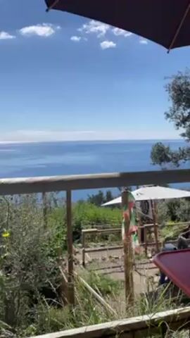 The video presents a scenic view of the sea under a blue sky, as seen from a high vantage point, likely a patio or balcony. The foreground is partially obstructed by a brown umbrella, and a wooden railing is visible. Below the railing, there's a garden area with plants, wooden fences, and white umbrellas shading tables and chairs. A man with a backpack walks by the railing.