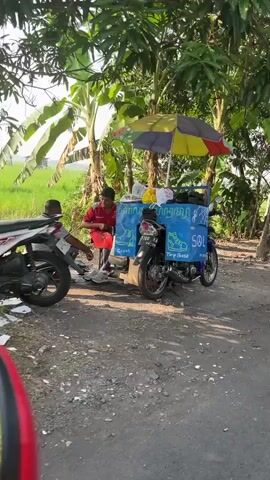 The video shows a street vendor selling food from a cart attached to a motorcycle. The cart is blue and has a colorful umbrella providing shade. There are two individuals present: a vendor in a red shirt serving a customer who is sitting on the back of another motorcycle. The vendor is handing something to the customer. The scene appears to be outdoors, with trees and what looks like a rice paddy in the background. The ground is dirt.