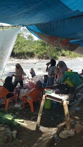 The video shows a group of people by a flowing river, likely in Meurah, Indonesia. The scene is set under makeshift shelters, one made of blue tarp and another of white fabric stretched over bamboo poles. Several individuals, including women in hijabs, are seated on orange plastic chairs near the water's edge, engaged in what appears to be a casual gathering or meal. One person is standing by a wooden table with various items on it. Another person is standing in the shallow water near the riverbank. The river is rocky with trees and lush greenery on the opposite bank, suggesting a natural, somewhat undeveloped setting. The lighting suggests it's daytime, possibly late morning or afternoon, with bright sunlight filtering through the trees. There are plastic bags and some debris visible on the ground, indicating human activity in the area. The overall mood seems relaxed and social, with people interacting with each other and enjoying the riverside environment.