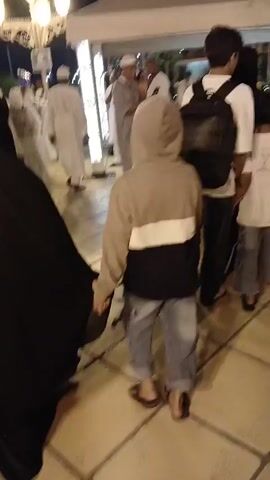 Here's a detailed description of the video:

The video shows a crowded outdoor scene at night. Several people are walking along a paved walkway. Some are dressed in traditional Islamic clothing, including a woman in a full black burqa and men in white thobes and head coverings. Other people are wearing casual clothing like t-shirts, jackets, and jeans. There is a child wearing a hoodie and jeans walking in the foreground.

In the background, there are white tents or canopies lit from within, suggesting some kind of event or gathering. Some people are standing around tables under the tents. Decorative streetlights illuminate the area. The architecture of a tall building can be seen in the background. The atmosphere appears busy and social.
