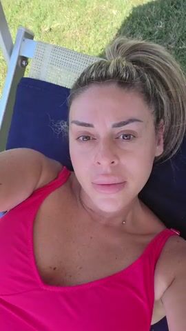 A woman with blonde hair pulled back in a ponytail lies on a lounge chair wearing a bright pink swimsuit. She is looking at the camera and her lips appear full. Her face is close to the camera. The background shows grass and the edge of the lounge chair. The lighting is bright and appears to be outdoors.