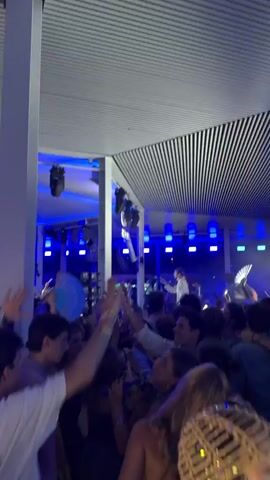 This video captures a lively concert or event taking place indoors, likely a venue or club. The lighting is dominated by vibrant blue hues cast from the ceiling, illuminating a large crowd of people. In the background, a band or musical performers are visible on a stage, equipped with speakers and lighting rigs. The audience is actively engaged, with many individuals raising their hands in the air, dancing, and appearing to enjoy the performance. Some people are holding up their phones, likely to record or take pictures. The overall atmosphere is energetic and festive.