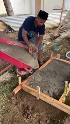 A man wearing a navy blue t-shirt, gray shorts, and a black taqiyah is pouring concrete from a red wheelbarrow into a square wooden frame on the ground. He is using a trowel to help guide the concrete, ensuring it fills the frame evenly. The frame is resting on soil and grass, and the surrounding area includes a building with a corrugated metal roof and a few scattered objects like buckets and hoses.