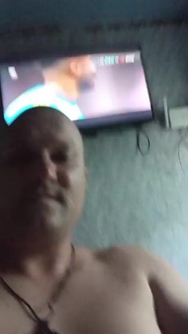 A man, bald and bare-chested, films himself from the chest up in a dimly lit room. He is looking directly at the camera and at one point holds up two fingers in a peace sign. Behind him, a television screen displays a sports game, possibly soccer, with players in uniforms. The wall behind the TV is covered in textured wallpaper. The lighting appears to be from the television and an overhead light source. The overall impression is of someone casually watching television.