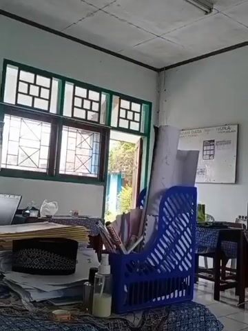 The video shows an interior view of a room, likely a classroom or office, with a focus on desks and a whiteboard. On the left side of the frame, a blue file organizer is filled with papers and documents. A bottle of hand sanitizer is also visible on a desk. In the background, a whiteboard displays text that appears to be a data chart. Two desks are set up with chairs, each covered with a blue and white patterned cloth. On the right side, a woman wearing a hijab is seated at one of the desks, looking towards the camera. Another desk is visible further to the right, with a blue file organizer on it. The room has white walls, a tiled floor, and a drop ceiling with fluorescent lighting. A window with green trim and square panes is visible on the left, and outside, trees and foliage can be seen. The overall impression is of a functional workspace.