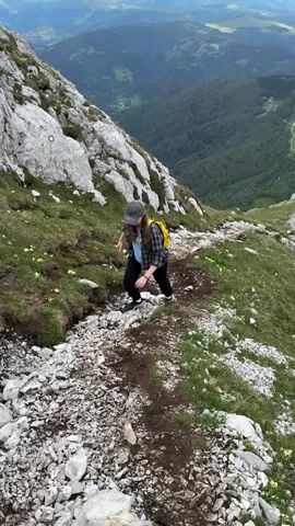 A person is hiking on a rocky, mountainous trail with a steep drop-off to the right. The hiker is wearing a patterned cap, a plaid shirt over a light blue t-shirt, and dark pants. They are walking downhill on a path strewn with rocks and patches of green grass, with small yellow flowers scattered amongst the grass.

The background is a vast, rolling landscape of green, forested mountains under a partly cloudy sky. A winding road is visible in the distance. The scene appears to be from a daytime hike. The overall mood is one of adventure and appreciation of nature. There is a red and white trail marker visible on a rock to the left of the frame.