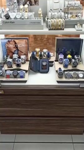 The video shows a display case filled with watches and jewelry. The top shelf features Calvin Klein watches and jewelry, as well as a display of jewelry with a focus on necklaces and bracelets. Below this, there are Nautica watches displayed on individual stands. To the right of the Nautica watches, there is a collection of watches with colorful bands. The second shelf from the top displays Lacoste watches, with a large Lacoste watch prominently featured in the center. The bottom shelf appears to be part of a store interior, with clothing racks visible in the background. The display case is well-lit, and the products are neatly arranged, suggesting a retail environment.