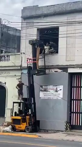 The video shows a yellow forklift lifting a black platform toward a window of a building in what appears to be a commercial or residential area. A man standing on the forklift seems to be giving instructions to several men who are inside the window. The platform carries a large rectangular object wrapped in brown paper and held in place with red straps. A sign on a nearby building reads "Maderas Belgrano..." in Spanish, suggesting this might be a furniture or woodworking business. The sky is overcast, indicating it's likely a cloudy day. The scene suggests a risky operation due to the man standing on the forklift and the precarious nature of the lift.