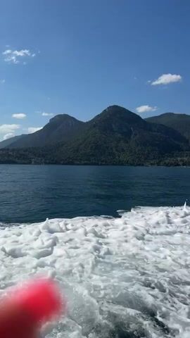 A video taken from a boat shows the wake of the boat in the foreground. The water is dark blue. In the background, green, tree-covered mountains rise up from the water. The sky is a clear, bright blue with a few white clouds. The sun is shining, creating a bright and serene atmosphere. The video is taken from a first-person perspective, giving the viewer the impression of being on the boat themselves.