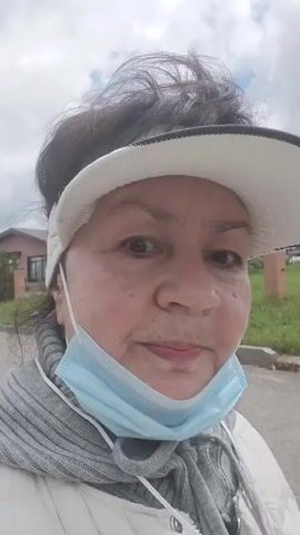 A medium close-up shot shows a middle-aged woman with dark, curly hair pulled back and wearing a white visor. She has a light blue disposable face mask hanging below her nose and a gray scarf around her neck. Her skin is fair with some freckles. In the first two seconds, she is sticking out her tongue and has a playful expression. In the last two seconds, she smiles broadly at the camera, her eyes crinkling at the corners. The background is slightly blurred, showing a house on the left and green grass and trees on the right under a cloudy sky. The lighting is natural daylight.